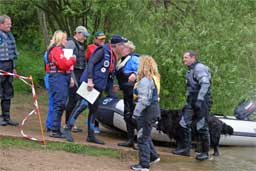 picture of group of people on lake shore by a boat, in the centre of the group a woman is being given a conratulatory kiss